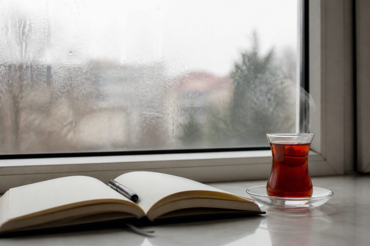 Buğulanmış pencere kenarında açık duran bir ajanda, kalem ve buharı tüten çay bardağı.